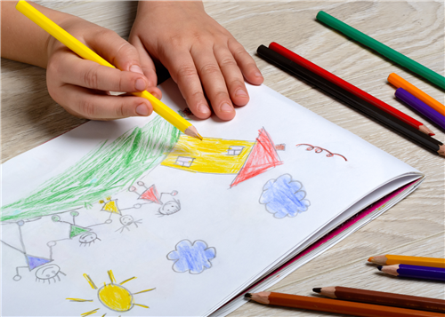 Closeup of child's hands holding a yellow coloring pencil, coloring a picture that includes a house, four members of a family